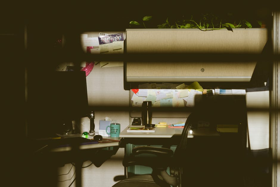 A dimly lit office cubicle featuring a computer, chair, desk, and indoor plants, illuminated by soft light.