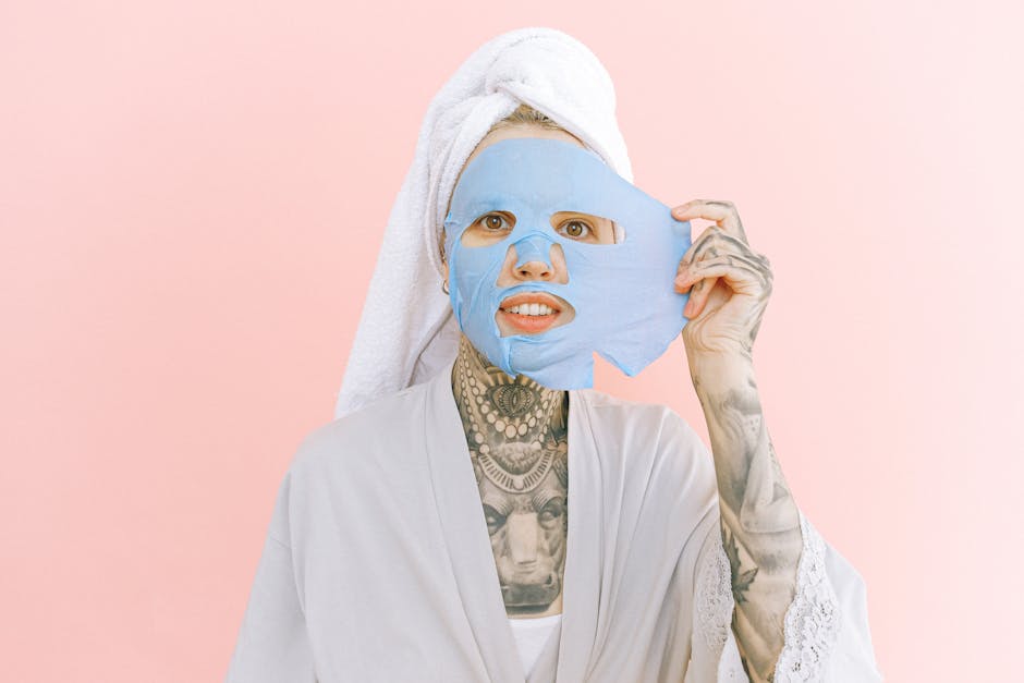 Woman in a white robe taking off a blue face mask with a pink background.