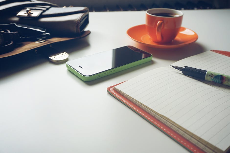 A tidy workspace featuring a coffee cup, smartphone, and open notebook with a pen.
