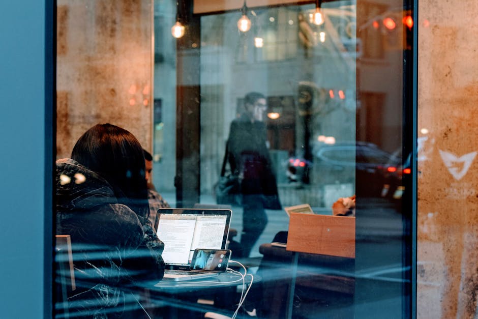 A person working on a laptop inside a café, viewed through a window with street reflections.