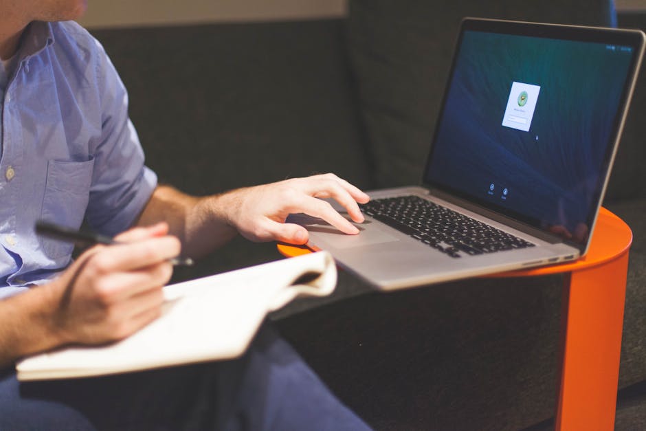 An adult man writing notes while working on a laptop indoors, illustrating a modern work environment.