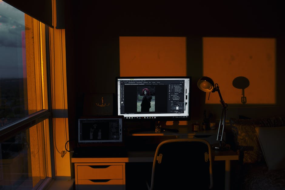 A dimly lit home office with a desktop and laptop illuminated by orange sunset light.
