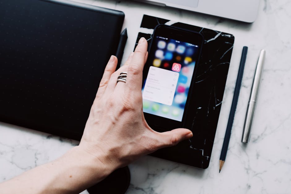 A hand holding a smartphone displaying apps on a marble desk, with a pen and pencil nearby.