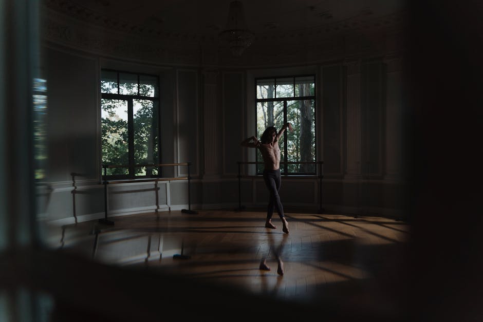 A shirtless dancer gracefully moves in a dimly lit studio, showcasing elegance and flexibility.