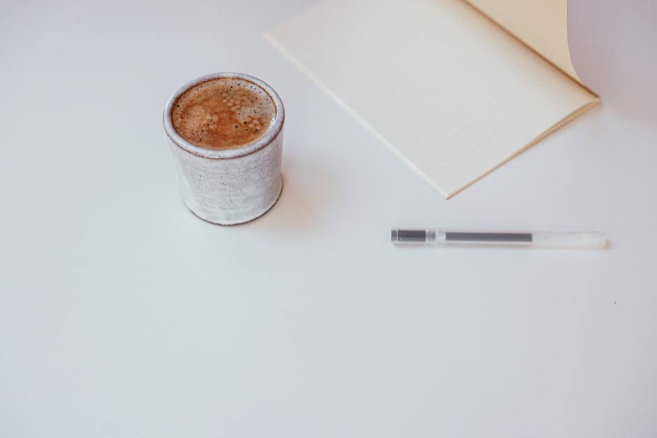 A minimalist workspace setup featuring a cup of coffee, notebook, and pen on a white surface.