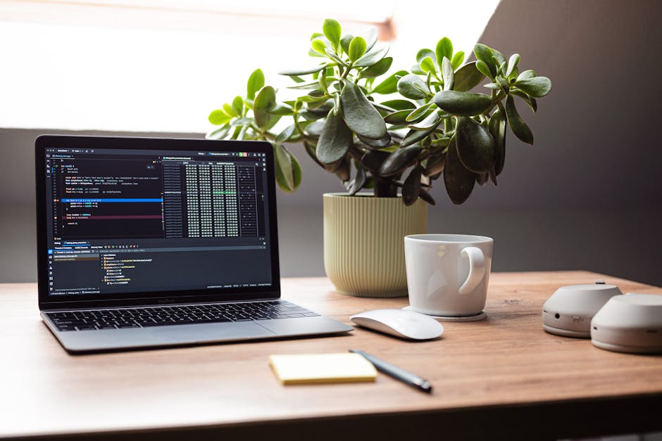 A modern workspace featuring a laptop, mouse, and plant on a wooden desk by a window.